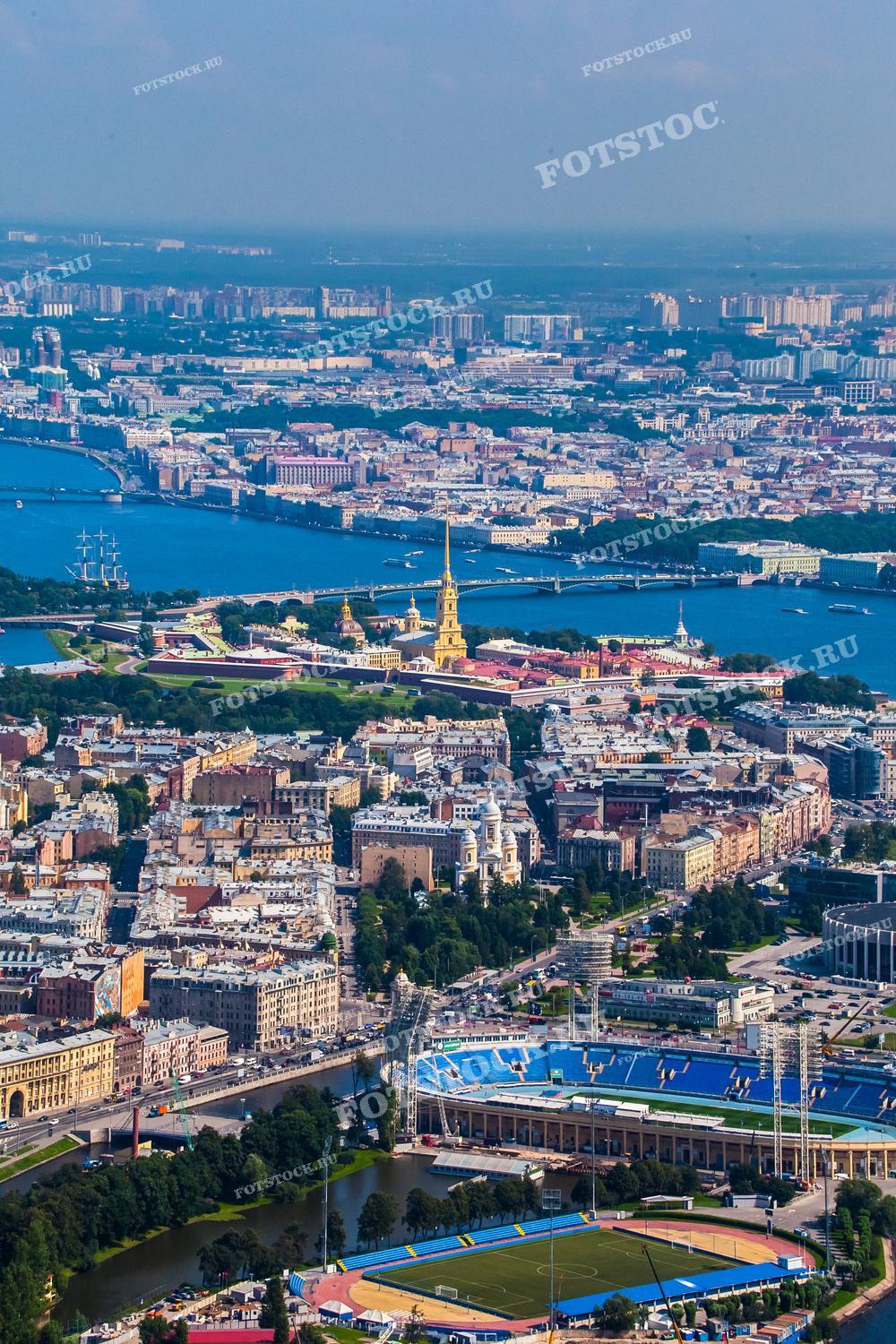 На фотографии изображена панорама Санкт-Петербурга с высоты. Виден Петропавловская крепость, река Нева и центральные районы города. Также можно увидеть спортивный стадион и множество исторических зданий. Фото может использоваться для туристических буклетов, открыток и рекламных материалов.